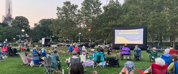 Wide shot of a large group of people sitting on colorful folding chairs on a grassy green lawn facing an outdoor projector screen that reads “Best Summer Ever.” The purple sky is turning to dusk in the background.