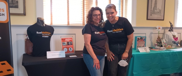 Two white women stand and smile in front of a table with ReelAbilities branded merchandise and flyers. The person on the right is Alison, a Humanities Engage fellow, and woman on the left is a Film Pittsburgh staff member. Both are wearing ReelAbilities t-shirts, jeans, and sneakers.